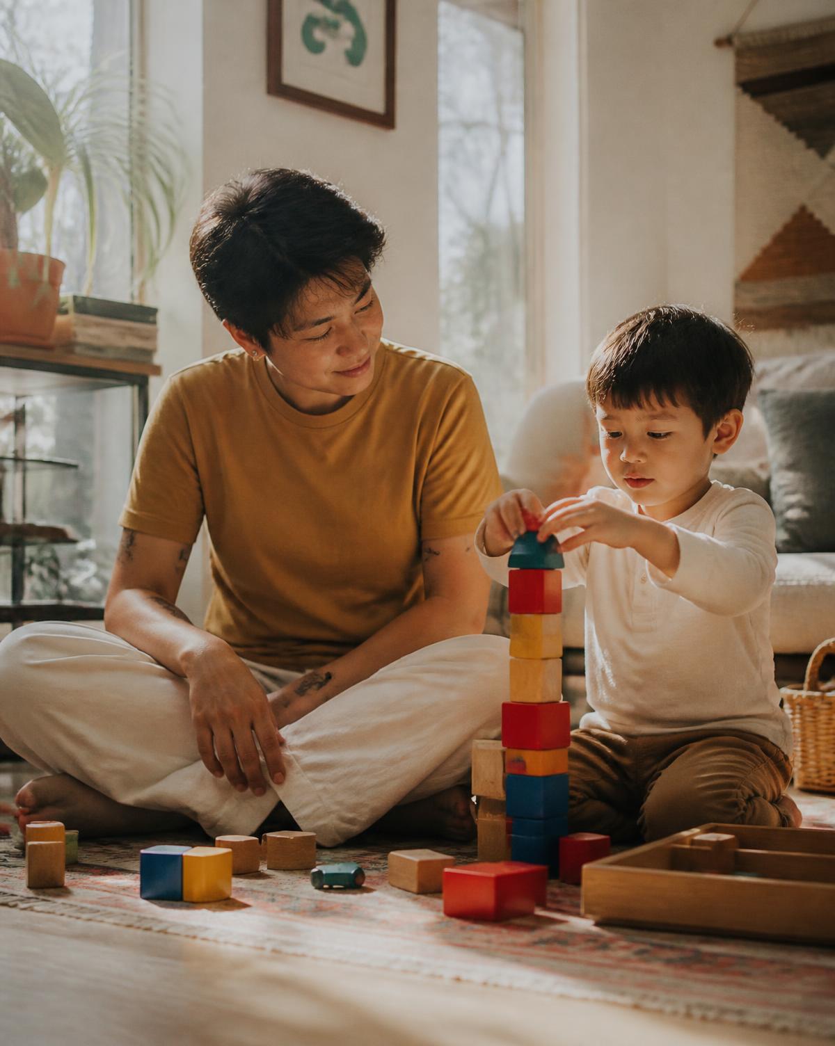 A parent sitting on the floor beside their child, who is stacking wooden blocks in a sunlit room